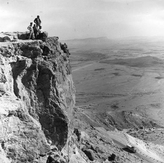 Hikers standing on the cliff edge above the Ramon Crater while a bus climbs the road below; a black-and-white photograph from the 1960s